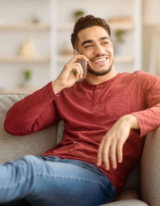 Happy handsome middle-eastern guy reclining on couch in cozy living room, talking on mobile phone with lover or friend, smiling and looking through the window, copy space, home interior